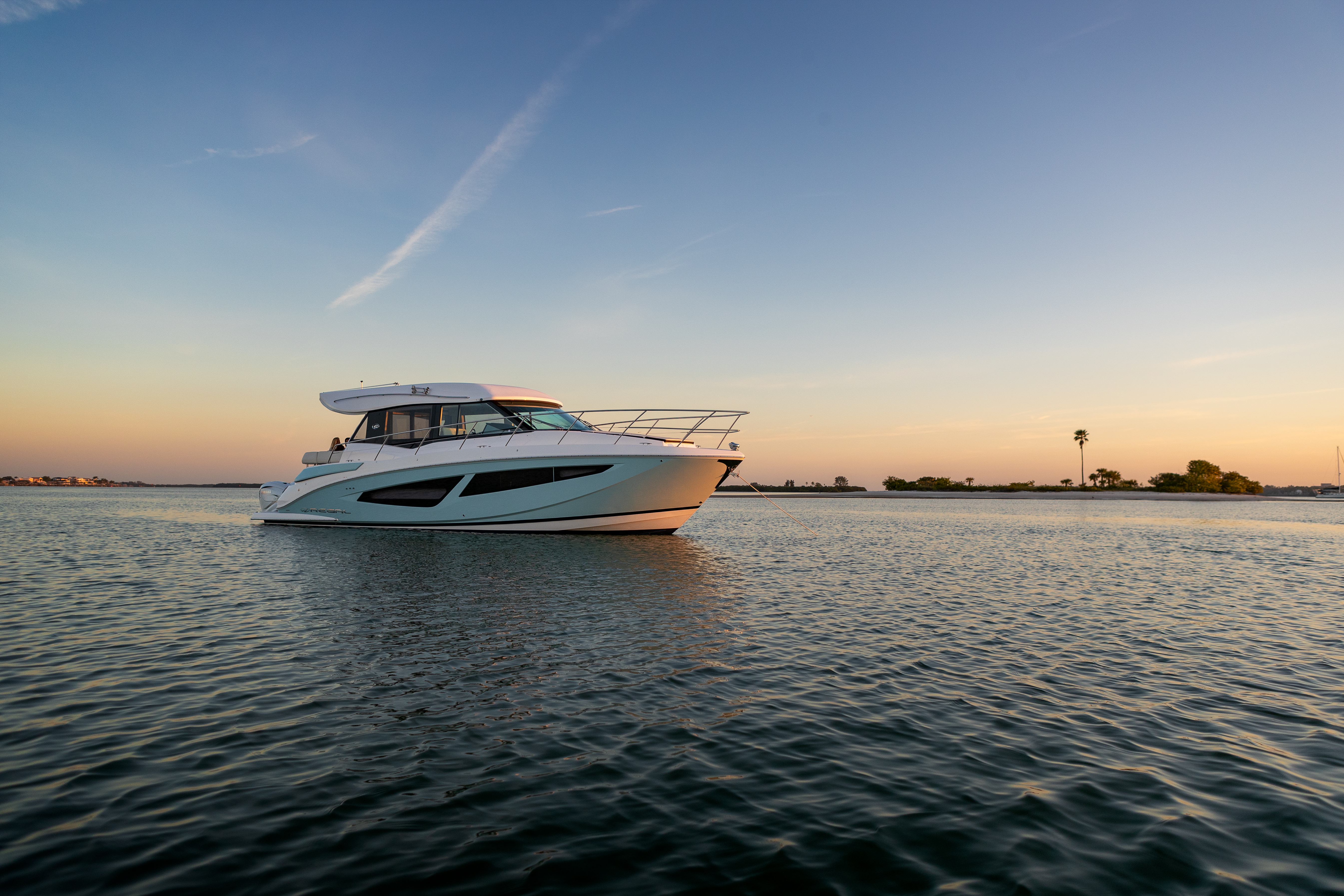 Regal luxury motor yacht anchored on calm water at sunset, with a low shoreline and palm trees in the distance.