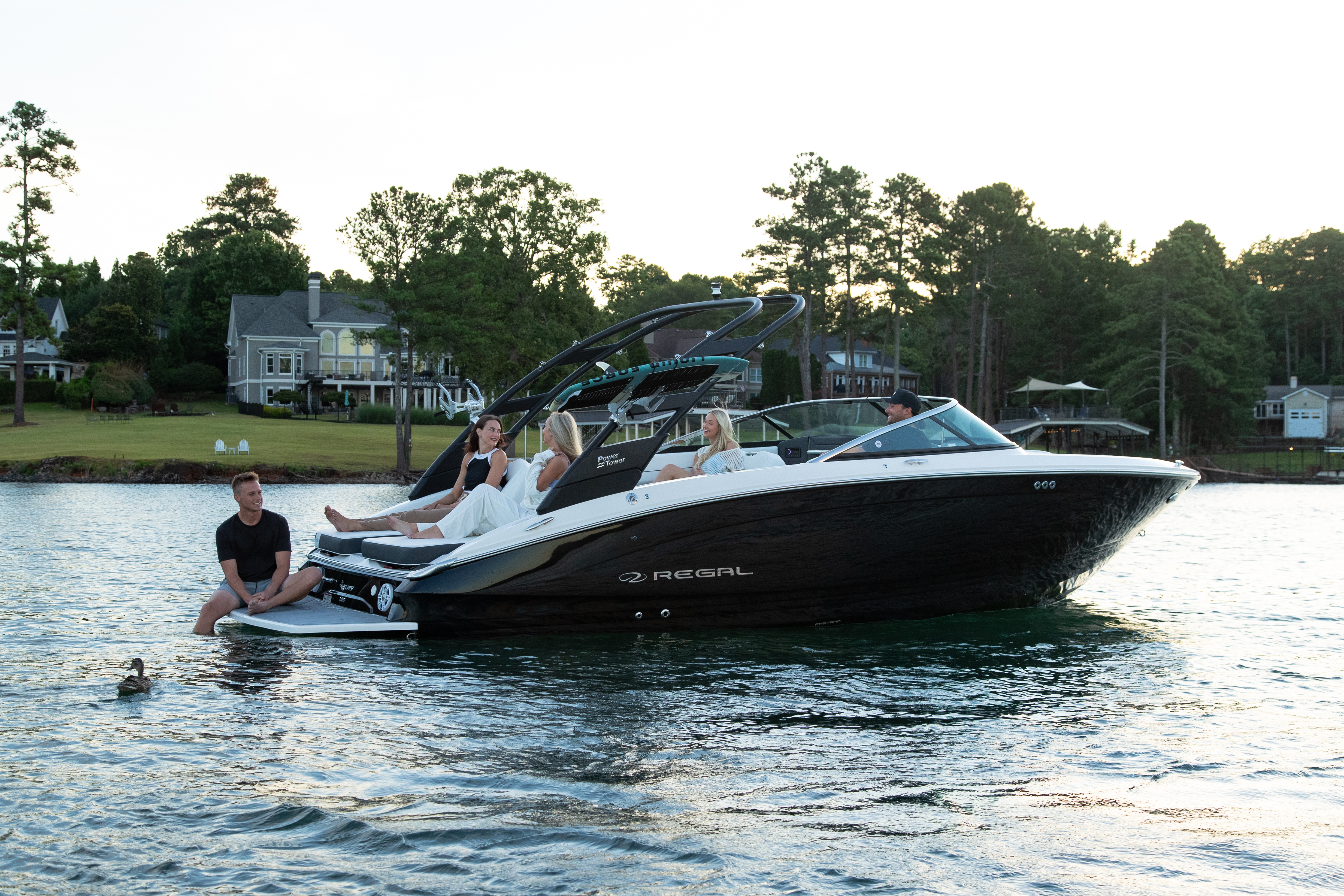 Regal LS6 Surf boat anchored near a waterfront neighborhood with five adults relaxing aboard and on the swim platform.